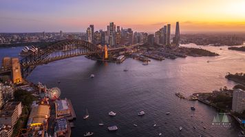 Aerial Sunset Over Sydney Harbour Bridge, Opera House and Crown Sydney Description: A breathtaking aerial photograph of Sydney Harbour at sunset, showcasing the Sydney Harbour Bridge arching gracefully across the water, the iconic Opera House...
