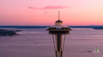 Aerial Seattle Sunset Space Needle and Bay A close-up view of Seattle’s Space Needle glowing at sunset above Puget Sound. Soft pink and lavender skies reflect across the calm evening water, with...