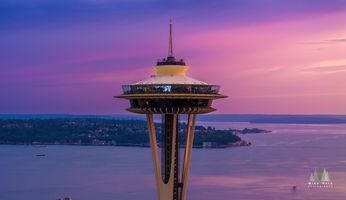 Aerial Seattle Sunset Space Needle An aerial close-up of Seattle’s Space Needle at sunset with Alki Beach and West Seattle visible beyond. The sky glows in brilliant tones of pink, purple, and...