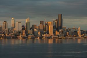 Seattle Dusk Waterfront Skyline from Alki Downtown Seattle skyline glowing in golden evening light with reflections across Elliott Bay under dramatic clouds.