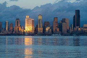 Seattle Skyline Brilliant Dusk Reflection Seattle skyline at sunset with the sun reflecting brightly off downtown skyscrapers above Elliott Bay under dramatic clouds.