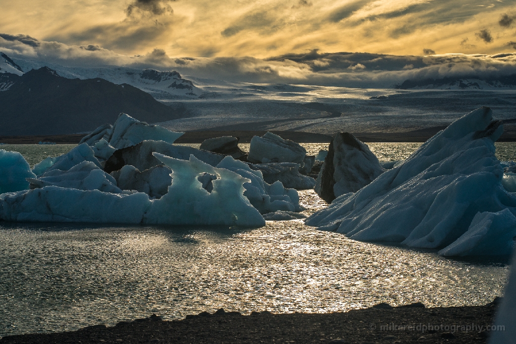 Sunlit Icebergs and Glacier Light at Jökulsárlón Iceland Warm sunlight glows across the surface of Jökulsárlón Glacier Lagoon, illuminating sculpted blue icebergs against a dramatic sky. The interplay of gold and turquoise hues captures the fleeting beauty of Iceland’s frozen landscapes. Fine-art photograph by Mike Reid revealing the balance of light, shadow, and texture in this Arctic wonder.