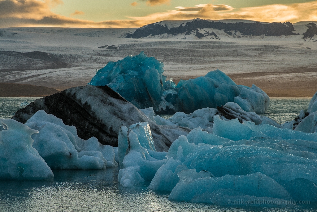 Blue Icebergs and Glacier Light at Jökulsárlón Iceland Brilliant blue icebergs drift across Iceland’s Jökulsárlón Glacier Lagoon beneath warm evening light and distant ice-covered peaks. The contrast between glowing glacier ice and golden sky reveals the fragile beauty of Iceland’s frozen landscapes. Fine-art photograph by Mike Reid capturing texture, light, and the serene power of the Arctic.