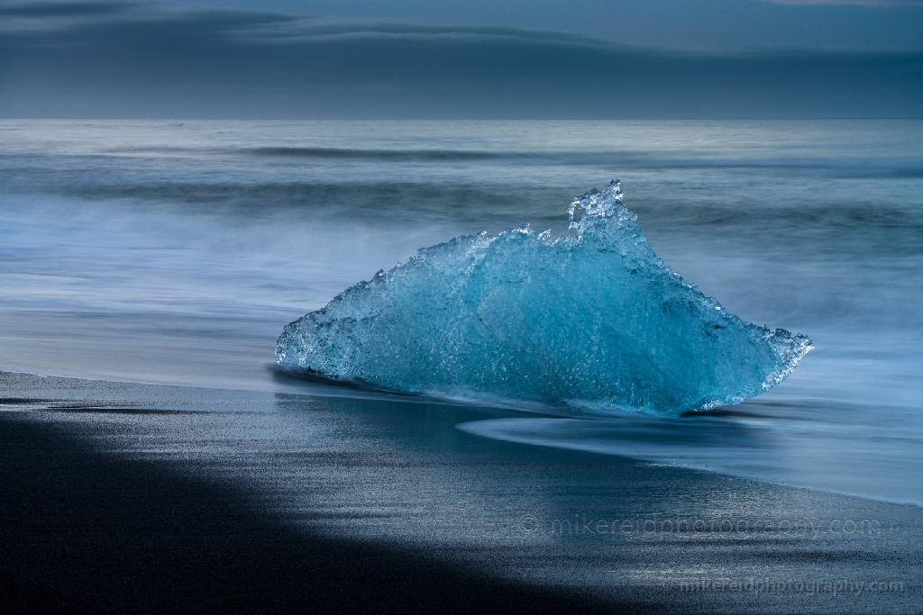 Blue Glacial Ice Closeup – Diamond Beach Jökulsárlón Iceland A luminous piece of blue glacial ice rests along the black volcanic sands of Diamond Beach near Jökulsárlón Glacier Lagoon in Iceland. The soft motion of the tide and cool Arctic tones create a striking contrast between the vibrant ice and dark shoreline. Captured by Mike Reid Photography, this fine art closeup showcases the texture, color, and sculptural beauty of Iceland’s frozen coastline.