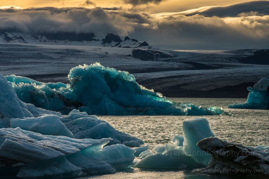 Iceland Jokulsarlon Sunset Hues of Blue Golden sunlight glimmers across the floating blue icebergs of Jökulsárlón Glacier Lagoon, captured with the Zeiss 100-300mm lens for exceptional clarity and tonal depth. The scene blends the icy stillness of the lagoon with the warm glow of the Arctic sun, creating a stunning contrast of light and texture. Fine-art photograph by Mike Reid showcasing Iceland’s glacial beauty through precision optics and artistic vision.