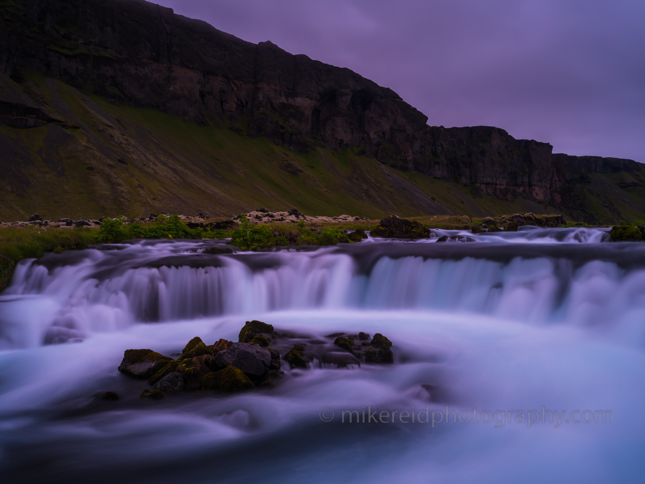 Iceland Ring Road Waterfall at Dusk – Serene Flow and Highland Cliffs Soft twilight light bathes a serene waterfall along Iceland’s Ring Road, where glacial waters cascade over dark volcanic rock beneath towering green cliffs. Captured by Mike Reid Photography, this tranquil long-exposure scene reveals Iceland’s ethereal beauty — silky motion, peaceful color tones, and the timeless contrast between earth and water. A fine art view of Iceland’s wild, meditative landscape.