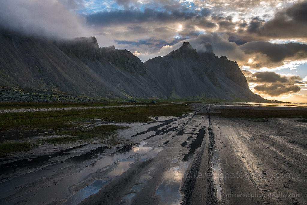 Iceland Road to Stokksnes Vestrahorn Low clouds drift across the dramatic slopes of Vestrahorn as early light filters through the stormy sky. Reflections shimmer in the wet black sands and tidal pools of Stokksnes Beach, revealing Iceland’s haunting beauty and rugged serenity. Fine-art landscape photograph by Mike Reid capturing mood, texture, and light on Iceland’s southeastern coast.