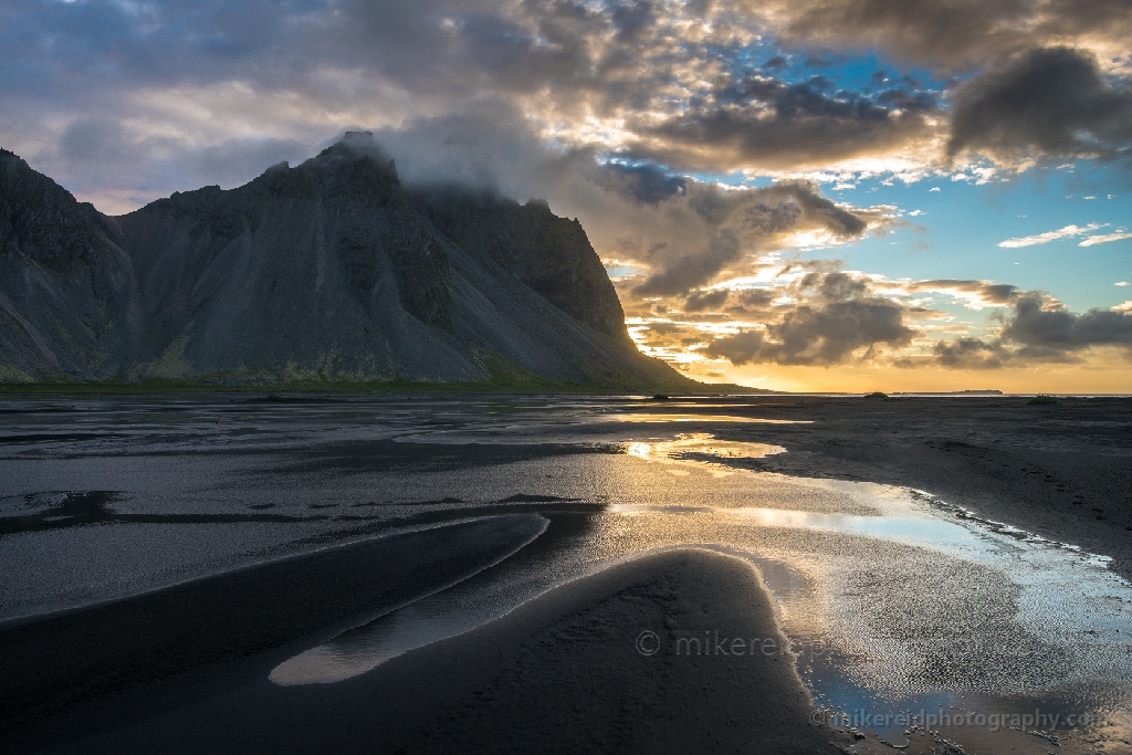 Iceland Stokksnes Vestrahorn Black Sands Reflection Soft morning light spills across the black sands of Stokksnes Beach, reflecting the rugged slopes of Vestrahorn under shifting clouds. The wet textures and golden tones evoke the raw beauty and solitude of Iceland’s southeastern coast. Fine-art landscape photograph by Mike Reid capturing the elemental light and atmosphere of Stokksnes.