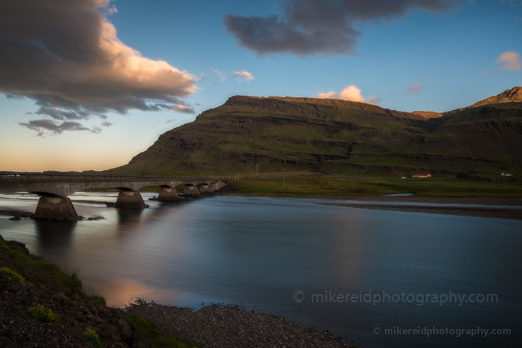 Evening Light and Water Motion beneath Ring Road Bridge Iceland A serene long-exposure photograph capturing the smooth motion of water flowing beneath an old Ring Road bridge in Iceland. The warm evening light glows against the rugged green hills and dramatic clouds, blending human architecture with untouched nature. Fine-art landscape photograph by Mike Reid, celebrating the timeless atmosphere of Iceland’s Ring Road and its quiet, scenic beauty.