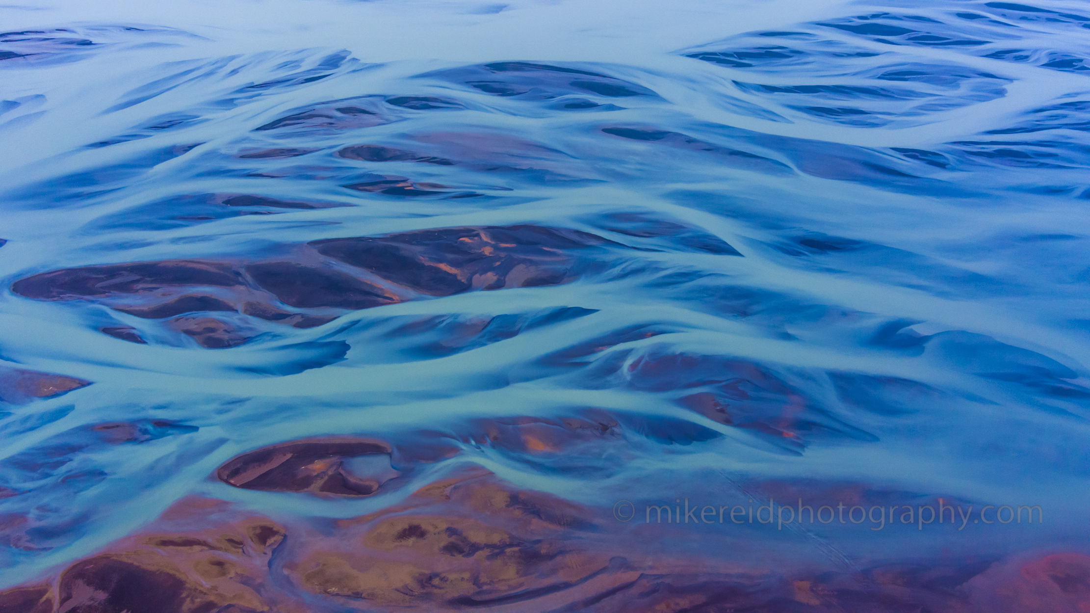 Iceland Braided River Aerial – Glacial Blues and Earth Tones From above, Iceland’s braided glacial rivers form an abstract tapestry of luminous blues and earthen purples, flowing gracefully across volcanic terrain. Captured by Mike Reid Photography, this aerial image transforms nature’s geometry into art — a mesmerizing study of color, light, and motion shaped by glacial meltwater. Perfect as a fine-art print for lovers of Iceland’s natural beauty and aerial abstraction.
