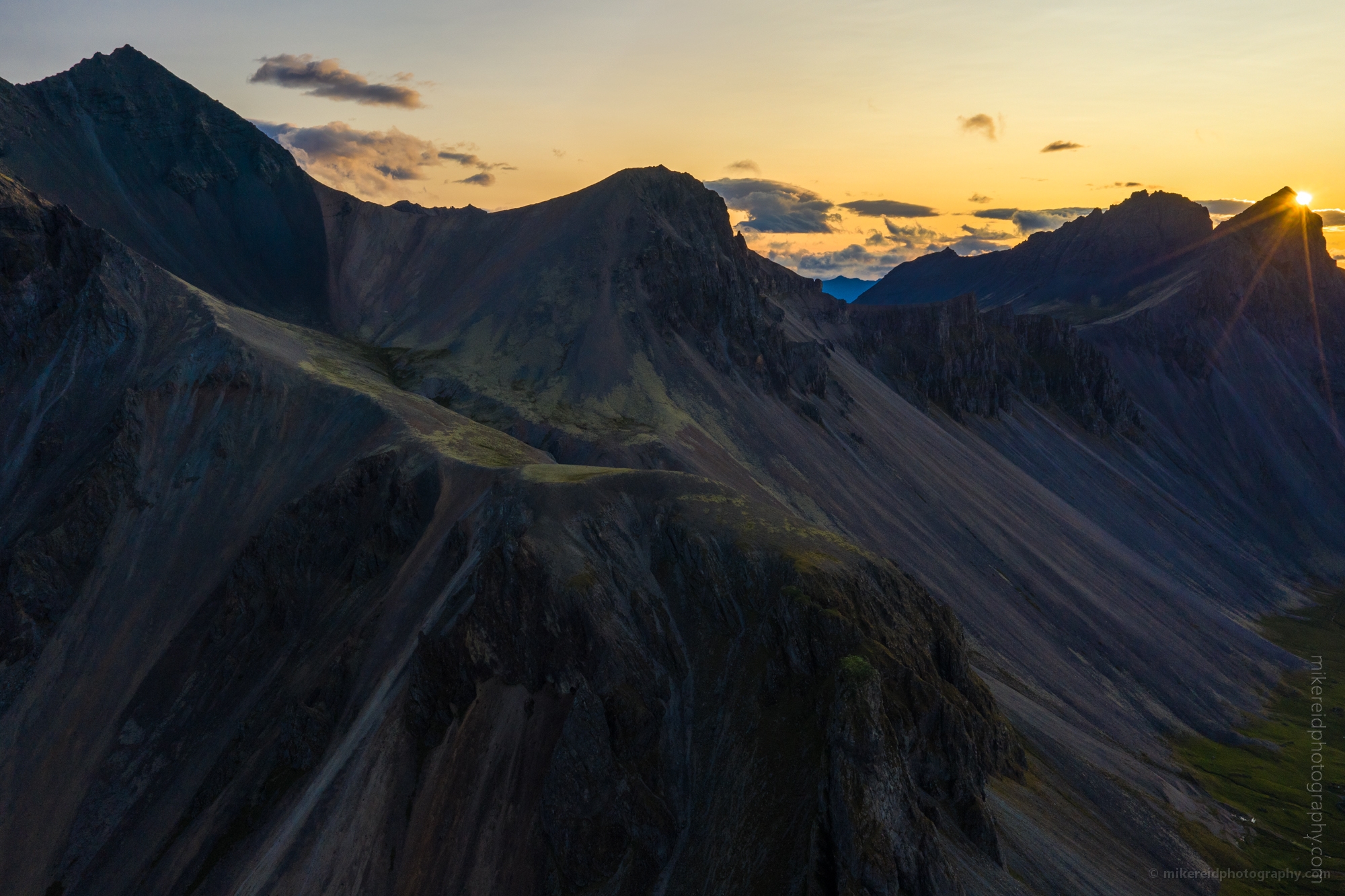 Sunrise Over Vestrahorn Peaks – Aerial View of Iceland’s Coastal Mountains The first light of dawn touches the sharp volcanic ridges of Vestrahorn Mountain, captured in this stunning aerial photograph above Iceland’s southeast coast. The golden sunrise spills across the rugged slopes, revealing layers of texture and shadow carved by time and weather. This fine art image embodies the wild spirit of Iceland’s landscapes — where light and land meet in breathtaking harmony.