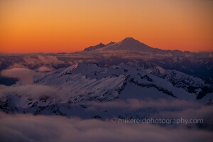 Over the North Cascades Mount Baker Sunset The last light of day paints Mount Baker and the surrounding North Cascades in glowing orange and violet tones as clouds drift among the peaks. Captured from...
