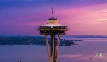 Aerial Seattle Sunset Space Needle An aerial close-up of Seattle’s Space Needle at sunset with Alki Beach and West Seattle visible beyond. The sky glows in brilliant tones of pink, purple, and...