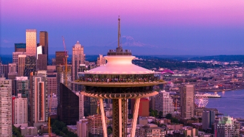 Aerial Space Needle Closeup A close-up aerial view of Seattle’s Space Needle glowing at sunset with downtown’s skyline and Mount Rainier in the distance. The city reflects soft pink and...