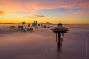 Over Seattle Space Needle and City Above the Clouds A dramatic sunrise over Seattle captures the Space Needle emerging above a thick blanket of fog with Mount Rainier glowing on the horizon. The downtown skyline...