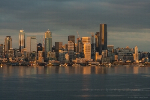 Seattle Dusk Waterfront Skyline from Alki Downtown Seattle skyline glowing in golden evening light with reflections across Elliott Bay under dramatic clouds.