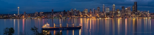 Seattle Skyline Night Reflection from Alki anoramic view of the Seattle skyline at blue hour from West Seattle, with the Space Needle and illuminated skyscrapers reflected across Elliott Bay as a cargo...
