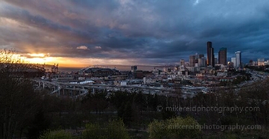 Sunset Over Seattle and Stadiums from Rizal Park A dramatic sunset unfolds over Seattle as seen from Rizal Park, with light breaking through stormy clouds to illuminate the skyline and stadiums below. The warm...