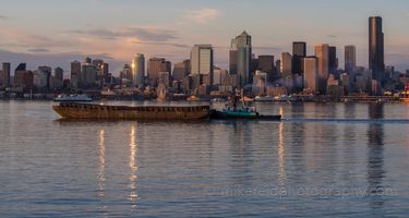 Seattle Photography Working Harbor at Dusk with Tug A tugboat and barge glide across the still waters of Elliott Bay under the golden light of sunrise. The Seattle skyline rises in the background, glowing with...