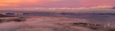 Pink Glow Over the Alaska Coast Range Soft pink and coral light spreads across the sky above Alaska’s Coast Range as low clouds and fog drift through the waterways below. Snow-covered peaks rise...