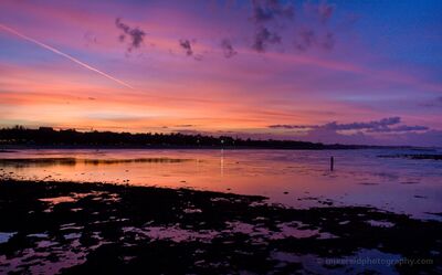 Bali Fisherman at Twilight A lone fisherman stands in the shallow coastal waters of Bali as vivid pink, orange, and violet twilight colors spread across the sky and reflect on the tidal...