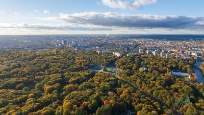 Tiergarten Autumn Aerial Berlin A sweeping aerial view over Berlin’s Tiergarten in autumn, with the park’s dense canopy turning shades of green, gold, and orange beneath a broad morning sky....