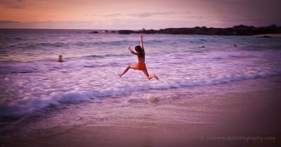Joy at the Edge of the Pacific A young traveler leaps into the surf at sunset on Hawaii’s Big Island, caught in a spontaneous moment of pure excitement. Soft pink and violet evening light...