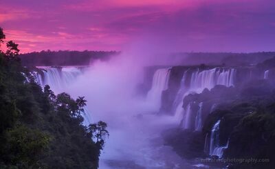 Iguazu Falls in Magenta Dawn Light A dramatic sunrise view across Iguazu Falls, with multiple cascades pouring through dense rainforest beneath an intense magenta and violet sky. Mist rises from...