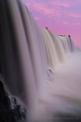 Iguazu Falls at Pink Sunrise A close sunrise view of Iguazu Falls, with a massive curtain of water dropping into thick mist beneath a vivid pink and violet sky. The soft dawn color...