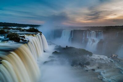 Iguazu Falls at Sunset A sweeping sunset view of Iguazu Falls, with powerful curtains of water dropping into heavy mist beneath a softly glowing evening sky. The nearest falls...
