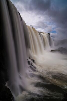 Iguazu Falls Under Dramatic Skies A dramatic close view of Iguazu Falls, with immense curtains of water plunging into heavy mist beneath dark, fast-moving clouds. A break of blue sky adds...