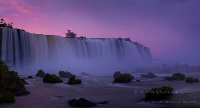 Iguazu Falls at Violet Dawn A panoramic dawn view of Iguazu Falls, with a broad curtain of water stretching across the frame beneath a vivid violet and pink sky. Mist rises through the...