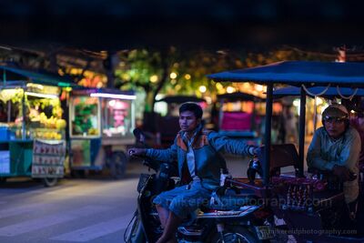 Tuk Tuk Drivers at Night in Siem Reap Two tuk tuk drivers wait beside the glowing street stalls of Siem Reap, Cambodia, after dark. Cool blue evening light mixes with warm market illumination,...