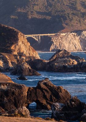 Bixby Bridge in the Distance Bixby Creek Bridge appears in the distance beyond rugged coastal rocks and sunlit cliffs along the Big Sur shoreline. Warm afternoon light and layered headlands...