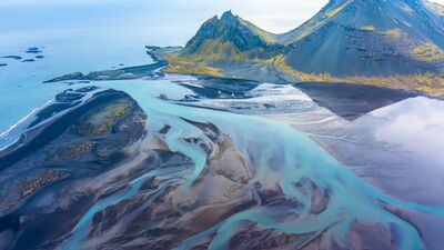 Aerial Stokksnes Estuary Flow – Iceland Coastline from Above An ethereal aerial photograph over the Stokksnes estuary in southeastern Iceland, where glacial meltwater fans into the sea beneath the dramatic peaks of...
