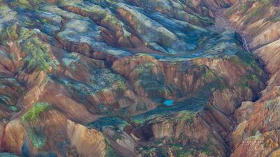 Hidden Blue Lake in the Rhyolite Mountains of Landmannalaugar An aerial view across the colorful rhyolite mountains of Landmannalaugar in Iceland’s Highlands, where bands of rust, green, blue-gray, and gold fold around a...