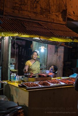 Street Food Vendor and Daughter in Chennai India A street food vendor works beside her daughter at a small illuminated stall in Chennai, India. Warm light spills across trays of food and the weathered...
