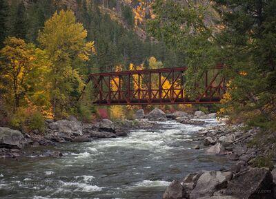 Autumn Wenatchee River Bridge A rust-red railroad bridge spans the Wenatchee River through Tumwater Canyon near Leavenworth, Washington, framed by brilliant autumn foliage and evergreen...