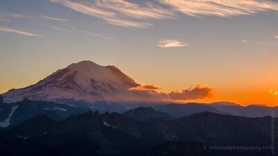 Mount Rainier Fiery Sunset Aerial — Pacific Northwest Mountain Photography An awe-inspiring aerial view of Mount Rainier silhouetted against a fiery Pacific Northwest sunset. Layers of alpine ridges fade into the distance as glowing...