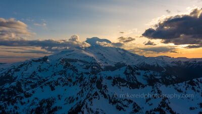 Over Mount Rainier Cowlitz Sunset Aerial Photography A dramatic aerial view of Mount Rainier at sunset, as golden light spills across the snow-covered ridges and peaks of the Cascade Mountains. Clouds swirl around...