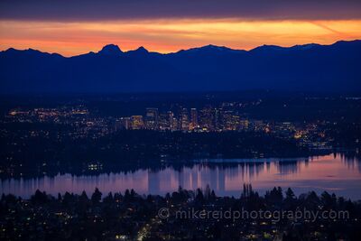 Bellevue Sunrise Beneath the Cascade Mountains A pre-dawn aerial view across Lake Washington toward downtown Bellevue, with the city lights glowing beneath the dark silhouette of the Cascade Mountains. Warm...