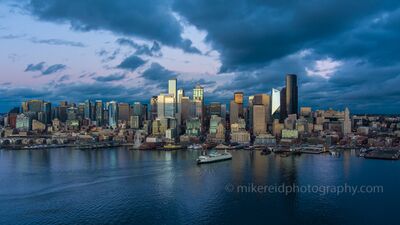 Over Seattle Ferry Arriving at Dusk An aerial view of downtown Seattle as evening settles in under dramatic storm clouds. The city’s glass towers reflect soft hues of fading daylight, while a...
