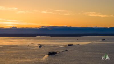 Tugboats Crossing Elliott Bay at Sunrise, Seattle Tugboats cross Elliott Bay beneath warm evening light as the Olympic Mountains fade into clouds beyond Seattle. This aerial view captures working vessels on...