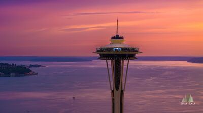 Aerial Seattle Sunset Space Needle Dusk An aerial view of Seattle’s Space Needle at sunset overlooking Puget Sound, bathed in soft hues of purple, pink, and orange. Sailboats drift below as the...