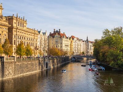Title Prague Waterfront in Autumn A warm autumn view along the Prague waterfront, with elegant historic buildings lining the river and trees turning gold and green along the opposite bank. Small...