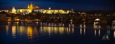 Prague Castle and Charles Bridge Reflections at Night A luminous nighttime view across the Vltava River toward Prague Castle and St. Vitus Cathedral, with the arches of Charles Bridge crossing the scene below. Warm...
