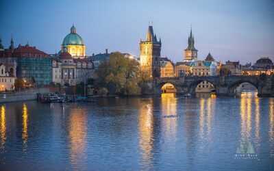 Charles Bridge Reflections at Dawn Prague A serene dawn view of the Charles Bridge and historic Prague riverfront, with warm architectural lights reflecting across the cool blue surface of the Vltava...