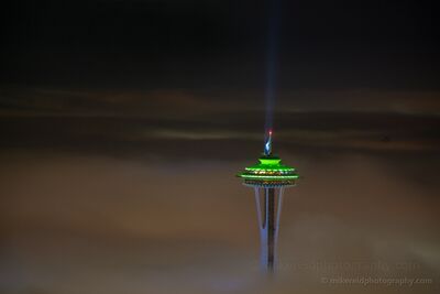Space Needle Above the Fog in Seahawks Colors The Space Needle rises above a dense layer of fog at night, illuminated in Seattle Seahawks blue and green. A narrow beam of light reaches into the dark sky...