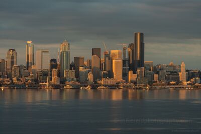 Seattle Dusk Waterfront Skyline from Alki Downtown Seattle skyline glowing in golden evening light with reflections across Elliott Bay under dramatic clouds.